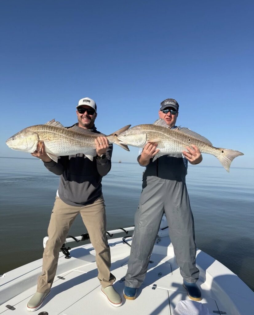 Captain Troy Wetzel and a guest each holding a large redfish on the bow of the boat on calm water