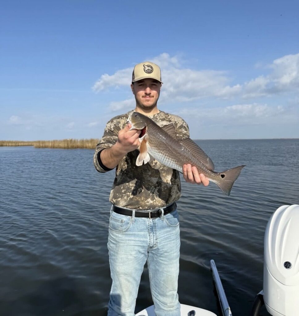 A guest on the bow of the boat holding a redfish, with marsh grass and open water behind